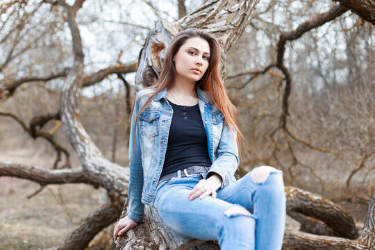 Stylish beautiful denim girl in fashion clothes with a jacket and torn jeans sits on a tree