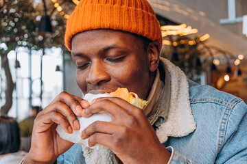 African American Man in Orange Beanie Savoring the First Bite of a Juicy Burger. A contented man takes a big bite of a burger, enjoying the meal amidst the cozy ambiance of a city eatery