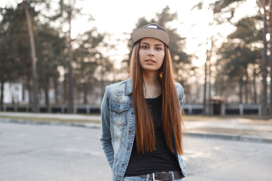 Urban beautiful denim woman in a stylish jacket and jeans with a black T-shirt with a cap walks on the street and looks at the camera