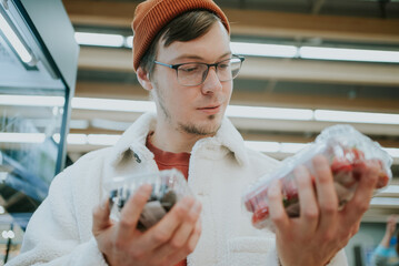 A thoughtful shopper compares containers of fresh berries, making a mindful selection of nutritious options in the produce section. Man Comparing Packs of Berries While Grocery Shopping 