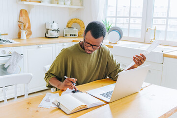 A focused African American man manages his finances at a kitchen table, using a laptop and calculator, with a receipt in hand, exemplifying personal accounting at home.
