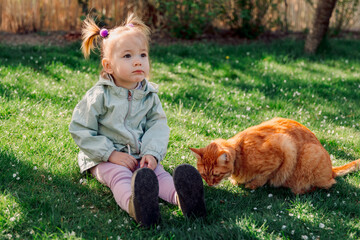 Child girl sitting on lawn grass with ginger cat in spring garden