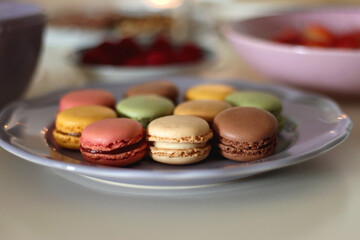 Plate of pastel macarons, cookies and chocolate, cup of tea of coffee, glass of bubble water, various berries, books and accessories on the table. Selective focus, pastel colors.