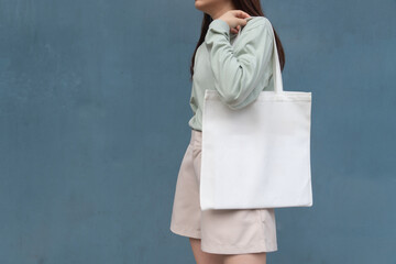 Blank white tote bag canvas fabric with handle mock up design. Close up of woman holding eco or reusable shopping bag against blue metal wall. No plastic bag and ecology concept.