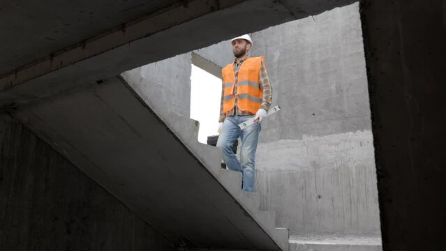 Builder repairman, foreman in protective helmet and vest stands at workplace in building and holds ruler and toolbox in hands