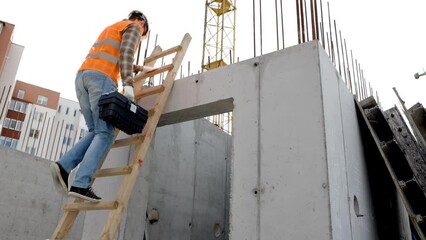 Maintenance worker man with safety helmet and orange vest climbing wood step ladder and holding toolbox at construction site