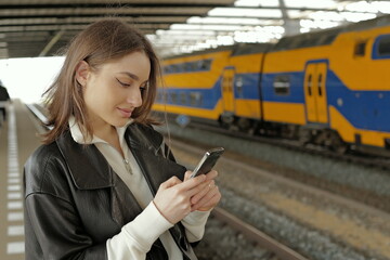 Portrait of a young woman using mobile phone waiting for a train at a station  - 781316995