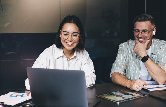 Happy male and female IT professionals reading publication about UI UX graphic design for web product and smiling at table desktop with modern laptop computer, cheerful colleagues collaborating