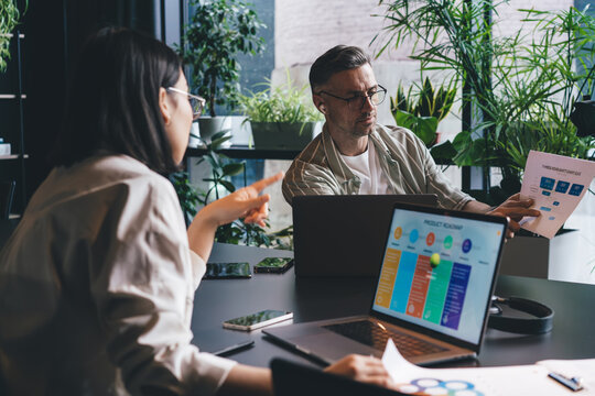 Male and female colleagues analyzing documents with infographics during cooperation brainstorming, skilled partners discussing business marketing during working process at desktop with laptops