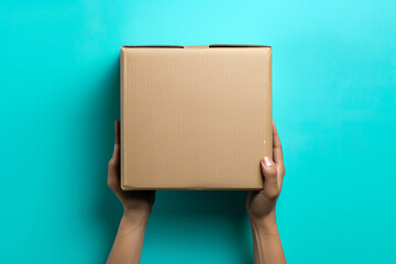 Top View of Hands Holding Cardboard Box. Overhead view of unrecognizable female hands holding a cardboard box on a turquoise background, ideal for shipping and delivery themes.