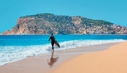 A male surfer walks on the beach with a surfboard in hand -  Landscape with marina and Red tower in...
