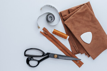 a stack of brown scraps of fabric lies on a white background, on top is a tailor's chalk, next to it are tailor's scissors, brown threads and a zipper, a measuring tape. shot from above