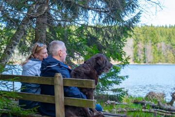 A couple of seniors with dog are sitting on the bench near the blue lake