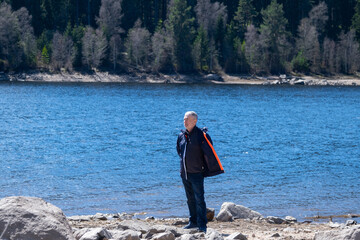 A senior man in jacket is staying on the bank near the blue lake