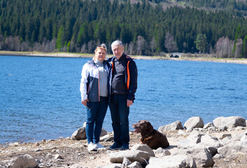 A couple of seniors with dog are staying on the bank near the blue lake