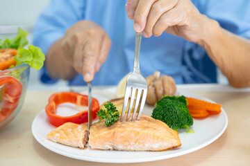 Asian senior woman patient eating Salmon steak breakfast with vegetable healthy food while sitting and hungry on bed in hospital.