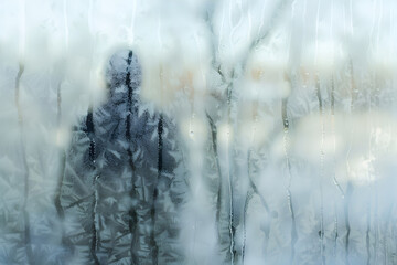 Silhouette of a person seen through a frosty window with intricate ice patterns and a cool blue winter atmosphere.