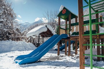 A playground stands empty on a snowy day, with a vibrant blue slide contrasting against the white...