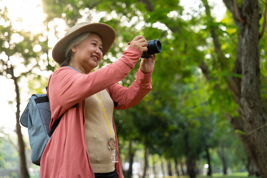 Portrait of Asian mature woman holding a camera and backpack behind her back, an Asia active senior woman enjoying nature in park. Standing on a trail in a forest outdoors. Enjoying active travel trip