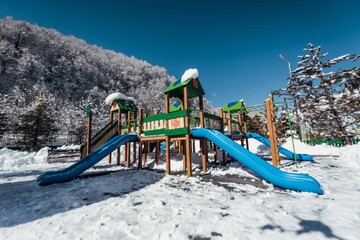 A playground stands empty on a snowy day, with a vibrant blue slide contrasting against the white...