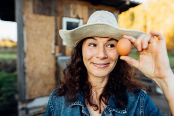 Portrait of funny joyful farmer woman holding fresh egg near her face. Portrait of a funny farmer with an egg in the summer. Happy moment of rural life