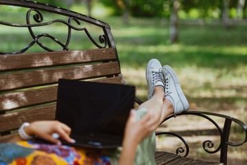 A woman works on her laptop while lying on a park bench, photo without a face. Freelancer lifestyle depicted by a woman working on technology in a quiet park