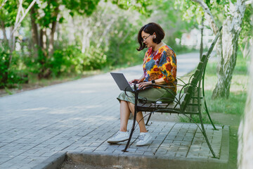 A cheerful woman works on her laptop while sitting on a park bench surrounded by birch trees. Casual businesswoman smiling as she successfully multitasks in urban park