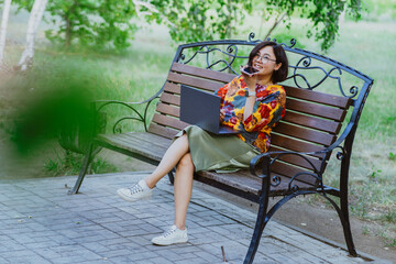 Smiling asian woman talking on speakerphone on smartphone while sitting with laptop on city park bench. Lifestyle freelancer, woman negotiating in park on smartphone outdoors in park.