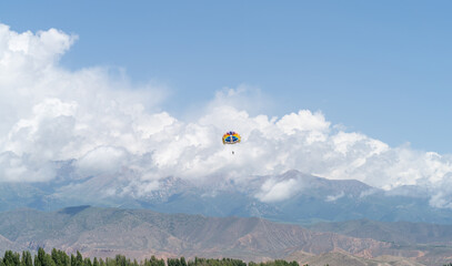 Two people are gliding on a colorful parachute against the backdrop of mountains and clouds in the...