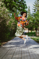 Asian adult Woman joyfully jumping with smartphone in sunlit park. Carefree youthful spirit captured with a Asian woman mid-leap