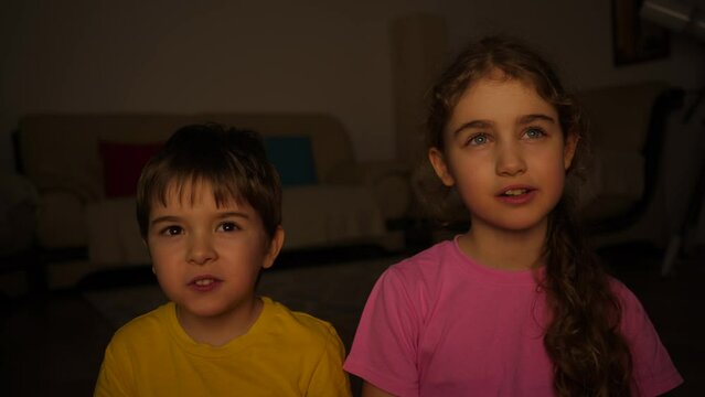 Children Watch Television at Evening. Brother and Sister Watching TV, Eating Popcorn Sitting on Floor at Home. Two Children Watching Exciting Movie in Living Room. Portrait Little Kids While Watch TV.