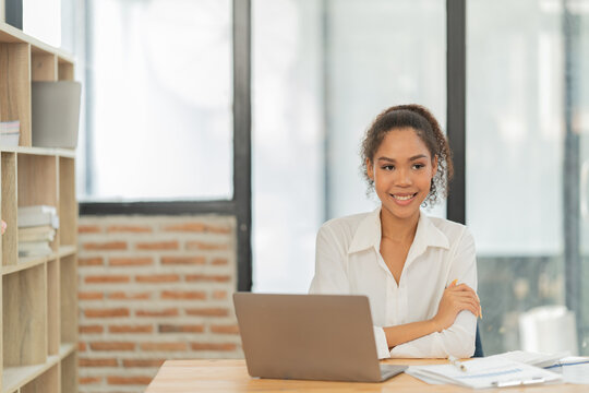 Young friendly businesswoman confidently looking away from her laptop, sitting at a wooden desk with documents and a bookshelf in the background, working in a modern office
