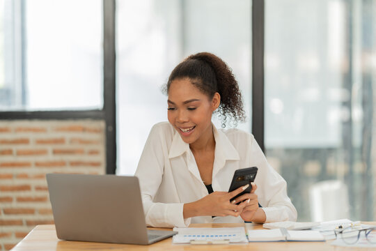 Professional woman working on laptop at her office desk, looking down at her smartphone and smiling, engaging in digital communication and multitasking with technology