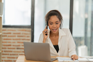 Young woman working in an office, using a laptop, speaking on a mobile phone, and reviewing documents on her desk, embodying productivity and communication in a modern workspace