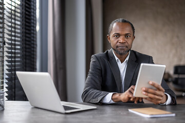 A man in a suit is sitting at a desk with two laptops and a tablet. He is focused on the tablet, possibly working on a project or browsing the internet. Concept of productivity and concentration