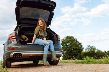 Young woman enjoying fresh air sitting in open trunk. Journey by car. Lifestyle, travel, tourism, nature concept.