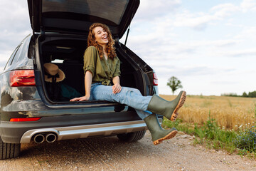 Young woman enjoying fresh air sitting in open trunk. Journey by car. Lifestyle, travel, tourism, nature concept.