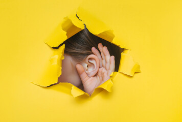 Female left ear and hand close-up. Copy space. Torn paper, yellow background. The concept of eavesdropping, espionage, gossip and the yellow press. - obrazy, fototapety, plakaty