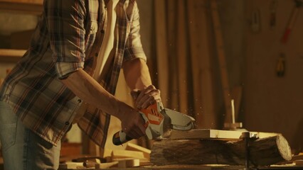 Male woodworker working in garage. Man professional carpenter working with wooden materials in workshop sawing wood using cordless mini chainsaw.