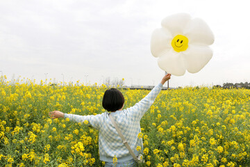 Back of a young woman as she holds a daisy balloon with a smiley face, wandering through rapeseed (Brassica napus) field full of bright colourful yellow flowers. Spring, summer, happy, joy concept