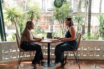 Two women enjoy collaborative work in tropical café. woman with braids smiles as she listens, while her companion gestures during conversation. laptop and note-taking indicate productive atmosphere - obrazy, fototapety, plakaty