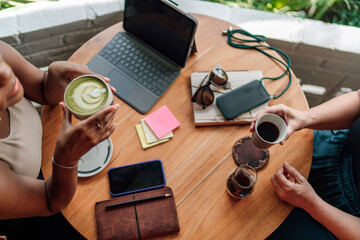 Two woman in casual meeting sharing moment over drinks, atmosphere suggests relaxed coffee shop setting with laptop computer and smartphones, indicating a blend of leisure and productivity..