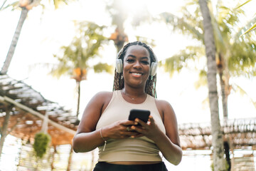 Smiling young African woman enjoying music on headphones, using smartphone in tropical setting,...