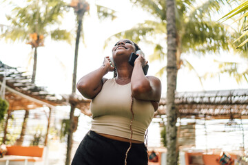 overjoyed African woman with headphones and smartphone, feeling the music, eyes closed, in tropical...