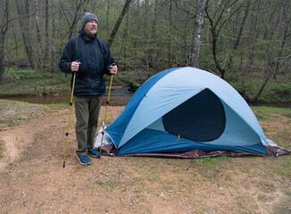 Senior hiker in cold weather with hiking poles standing by his tent.