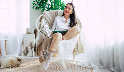 Happy lovely relaxed trendy woman with long healthy hair reads a book in stylish clothes in the modern living room while sitting on hanging chair.