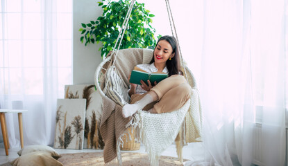 Happy lovely relaxed trendy woman with long healthy hair reads a book in stylish clothes in the modern living room while sitting on hanging chair. - 781407789