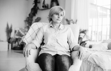 White and black photo of sad thoughtful mature woman looking away while sitting at home on the chair