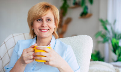 Happy lovely mid-aged woman resting with a smile while sitting with a cup of tea in her hands on the chair. Cheerful mature female enjoying a serene retirement at home.