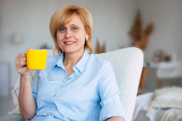 Happy lovely mid-aged woman resting with a smile while sitting with a cup of tea in her hands on the chair. Cheerful mature female enjoying a serene retirement at home.